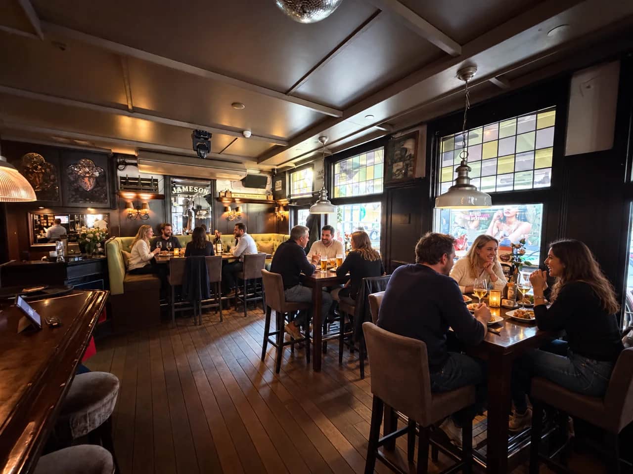 Gasten aan de hoge tafels naast de houten bar en glas-in-loodramen van De Appel